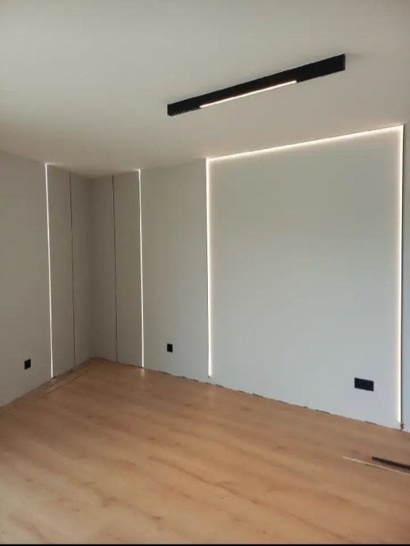 Pokój z drewnianą podłogą i nowoczesnym oświetleniem LED wzdłuż ścian i na suficie. Minimalistyczny design z czarnymi gniazdkami i listwami.