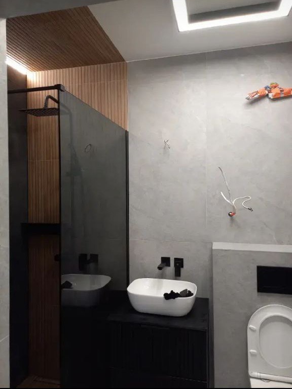 Nowoczesna łazienka z drewnianymi panelami, szarą płytką i białą ceramiką. Kabina prysznicowa typu walk-in, umywalka nablatowa i wisząca toaleta.