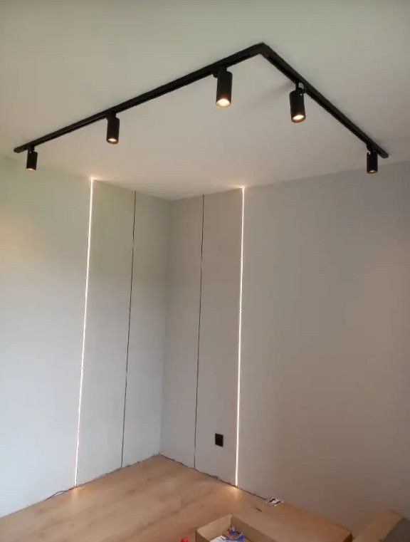 Nowoczesne oświetlenie sufitowe z reflektorami na szynie w rogu pokoju, minimalistyczne ściany z pionowymi szczelinami świetlnymi, parkiet na podłodze.