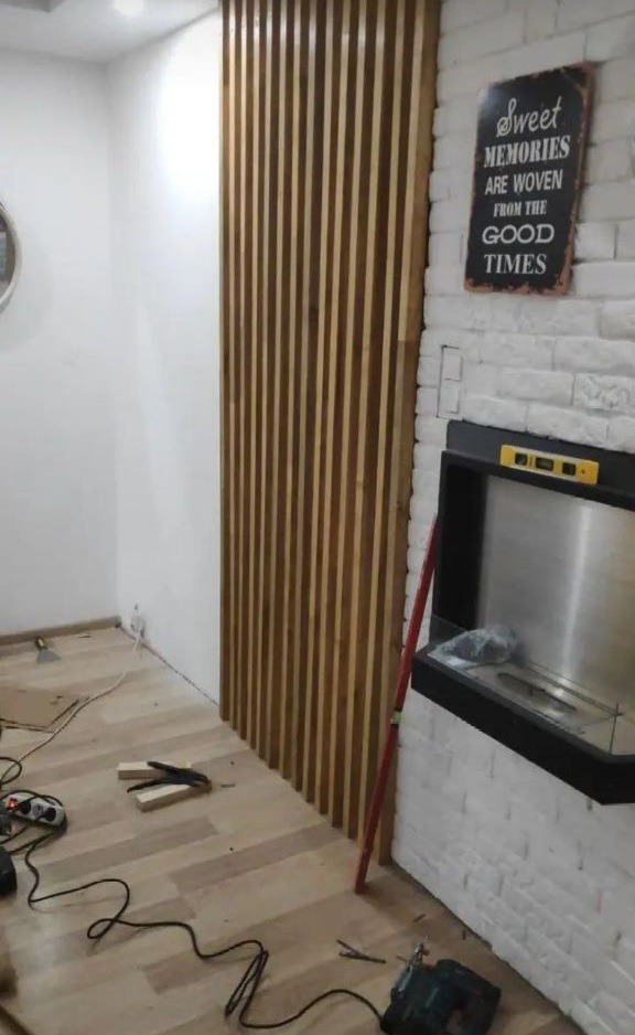 Drewniane lamele na ścianie, tworzące dekoracyjny panel. Widoczne narzędzia i poziomica, sugerujące montaż. Fragment kominka z cegły w tle.