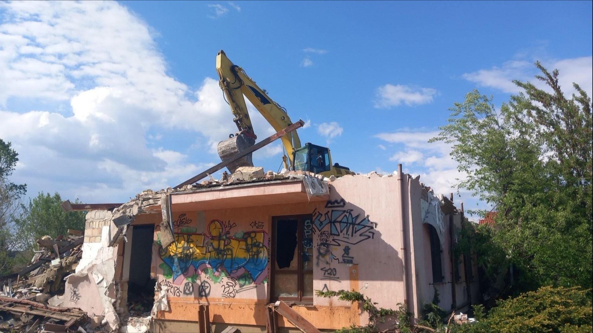 Żółty sprzęt ciężki na szczycie zniszczonego budynku z graffiti podczas rozbiórki w Prażmowie, błękitne niebo w tle.