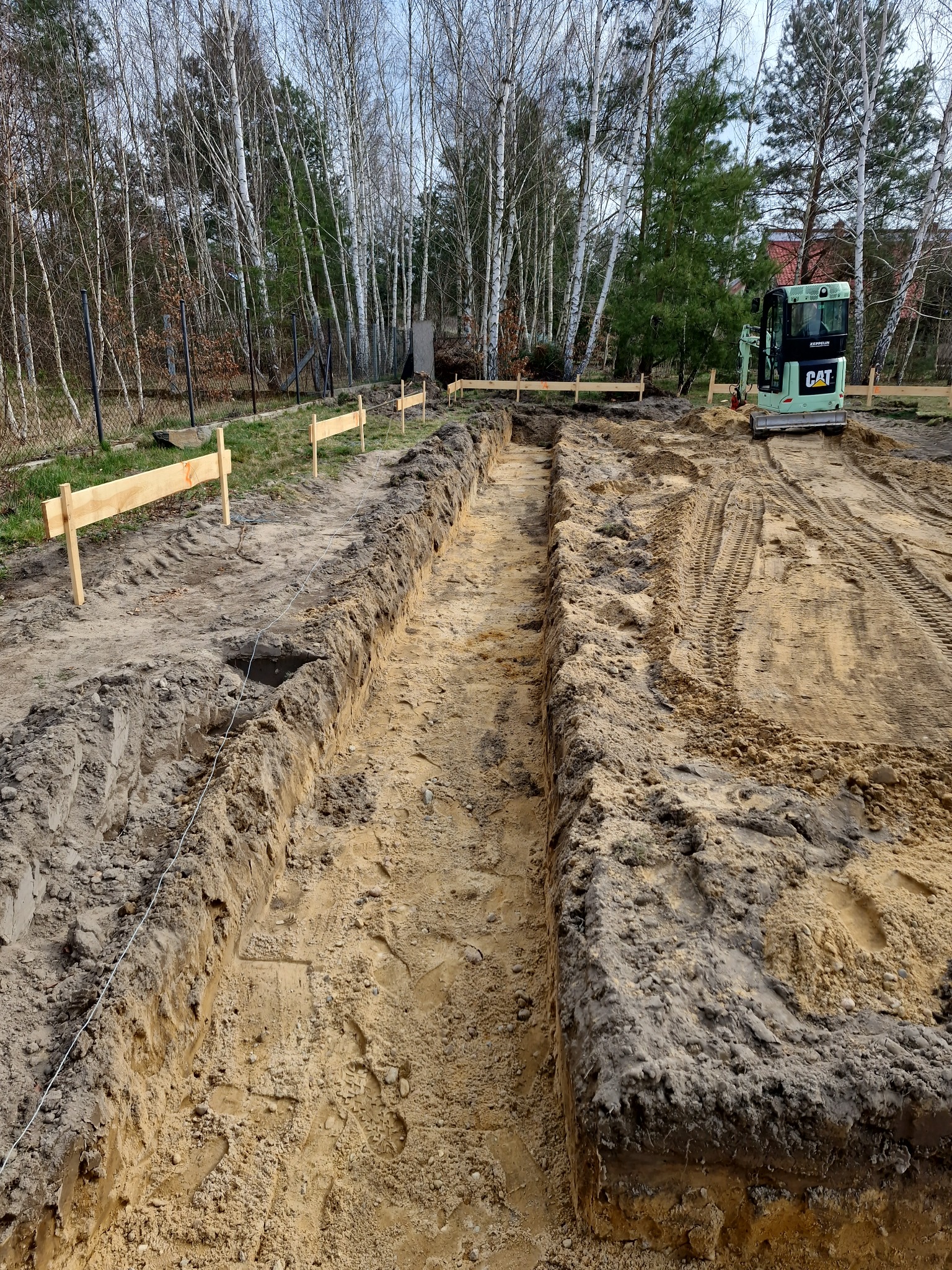 Wykopany wąski rów w ziemi pod fundament, wyznaczony drewnianymi słupkami i sznurkiem. W tle zielona mini-koparka CAT, drzewa i ogrodzenie.