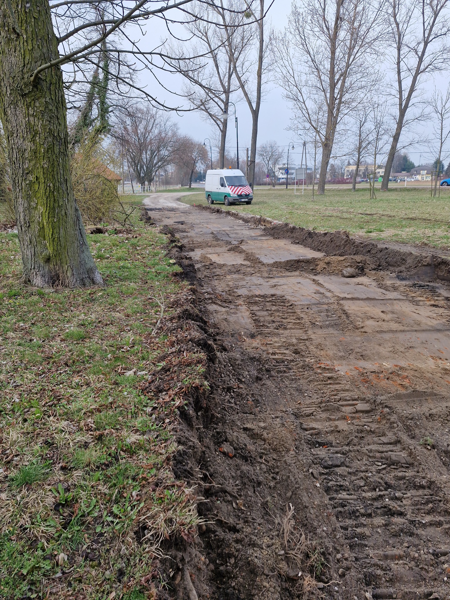 Przygotowanie terenu pod ścieżkę: wyrównany grunt z widocznymi śladami opon i wykopanymi brzegami. W tle park z drzewami i zaparkowany bus.