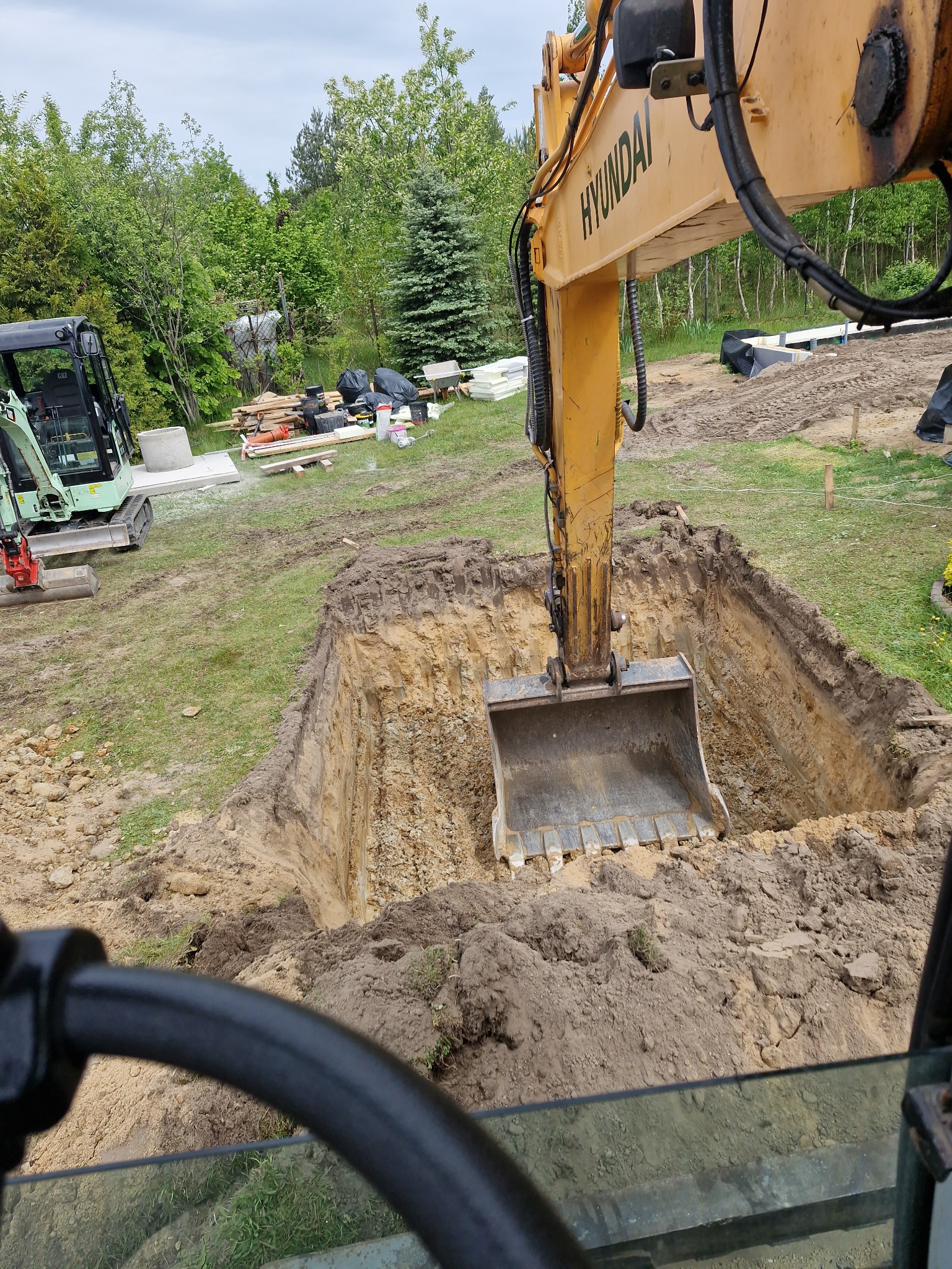 Żółta koparka Hyundai w trakcie wykopu prostokątnego dołu w ziemi, widok z kabiny operatora. W tle inny sprzęt budowlany i zieleń.