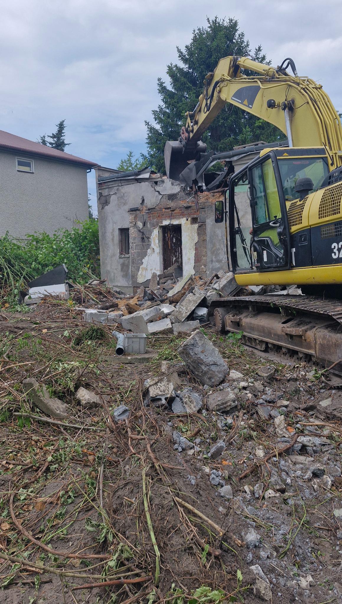 Żółta koparka z łyżką rozbiera murowany budynek. Gruz i resztki roślinności na ziemi. Fragment budynku sąsiada w tle. Dominuje pochmurna pogoda.