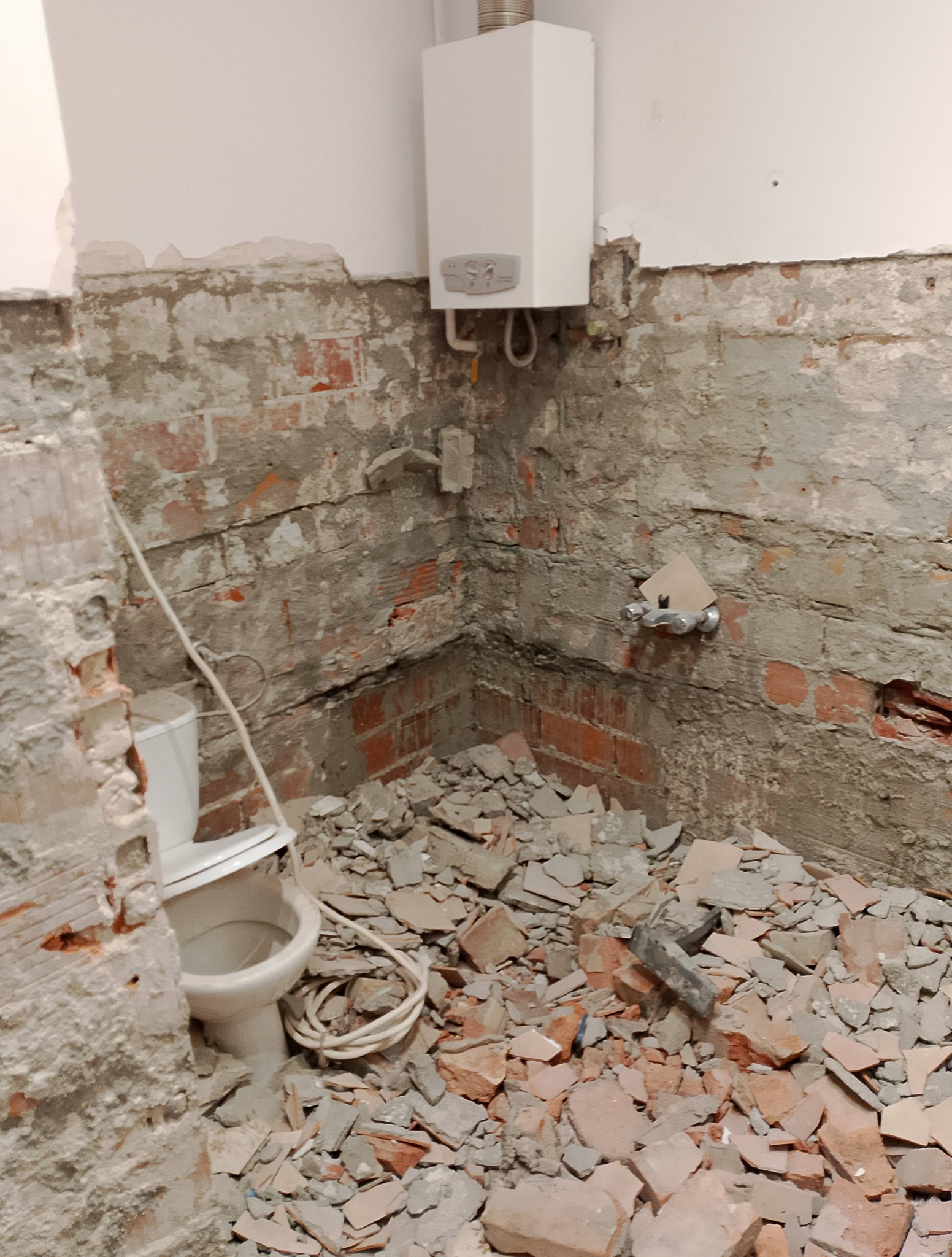 Demontaż płytek w łazience z odsłoniętą cegłą i wiszącym bojlerem. Widoczne gruz i fragment toalety. Etap prac remontowych przed wykończeniem.
