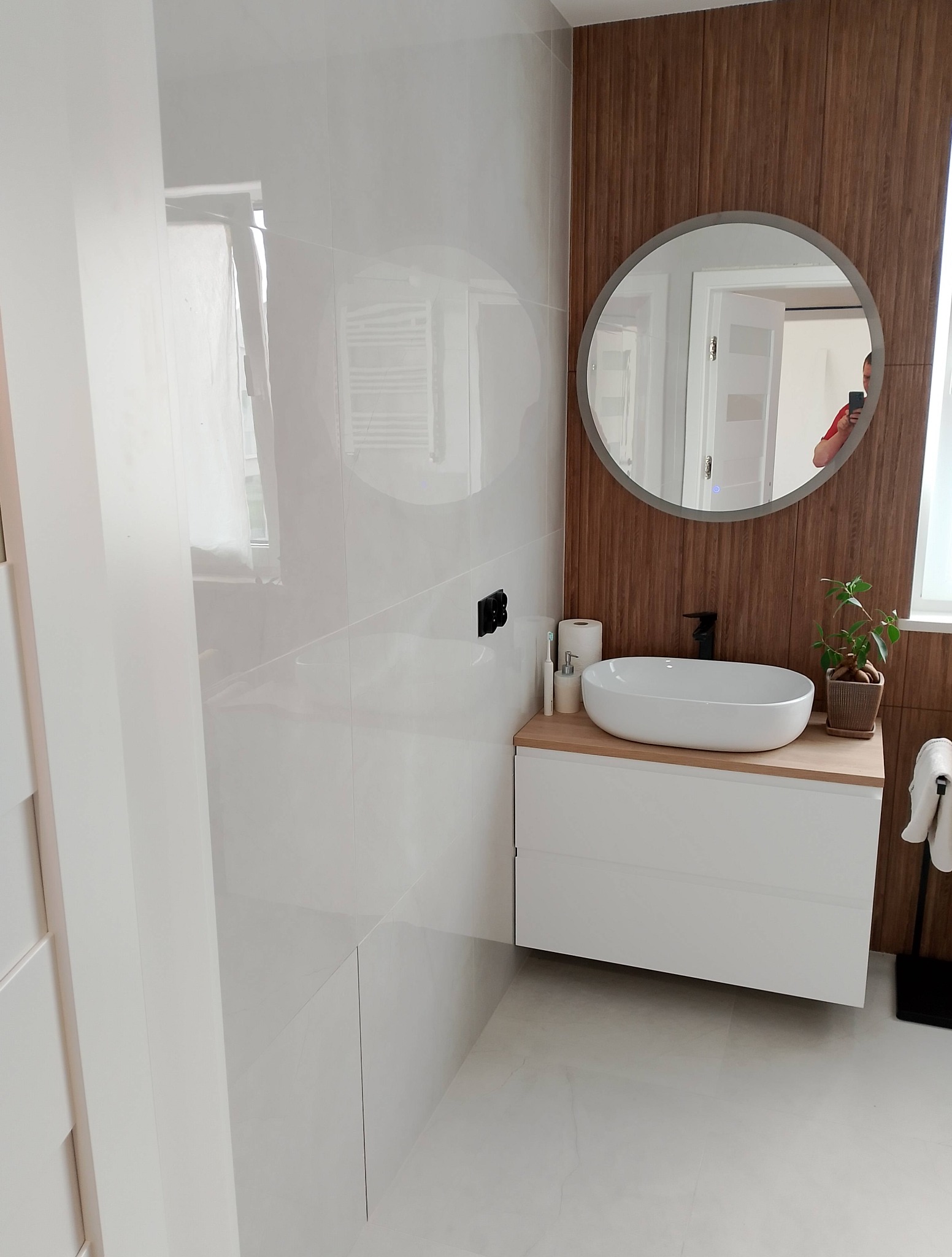 Nowoczesna łazienka z białymi płytkami, drewnianą ścianą i okrągłym lustrem. Umywalka nablatowa z czarną armaturą, akcent roślinny.