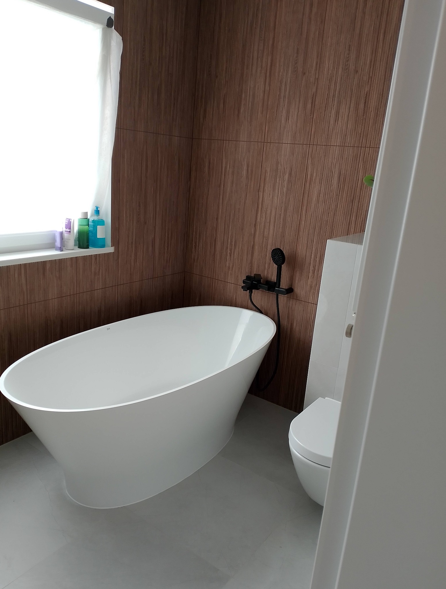 Nowoczesna łazienka z wolnostojącą wanną i drewnopodobnymi płytkami na ścianie. Czarna armatura dodaje elegancji. Okno z białymi zasłonami.