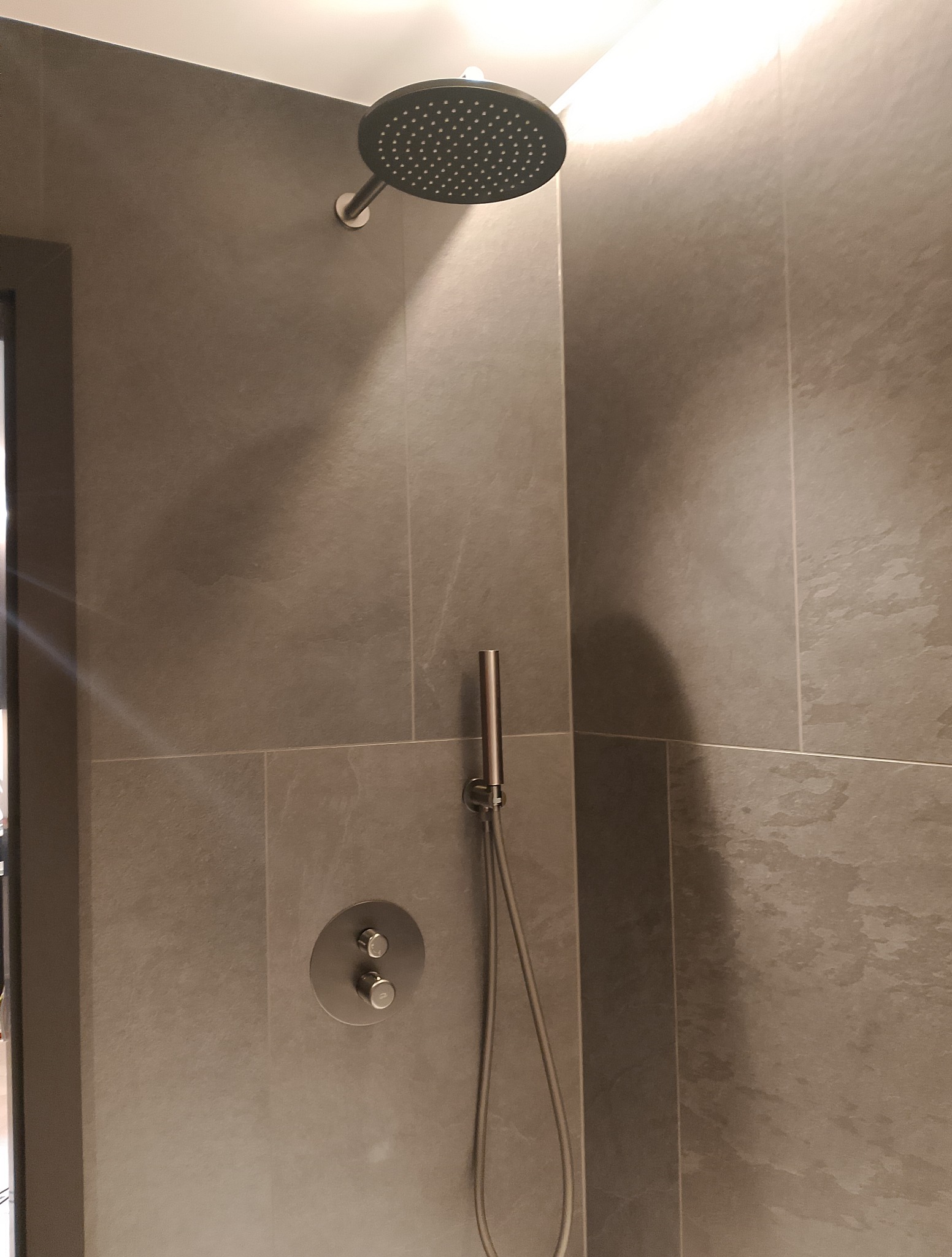 Nowoczesna łazienka z szarą, kamienną okładziną ścienną, deszczownicą i minimalistyczną baterią. Subtelne oświetlenie podkreśla teksturę płytek.