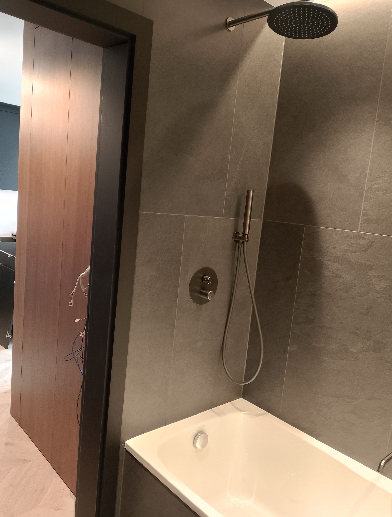 Nowoczesna łazienka z szarą, kamienną okładziną, deszczownicą i białą wanną. Widoczna armatura i fragment drewnianych drzwi z ciemną ościeżnicą.