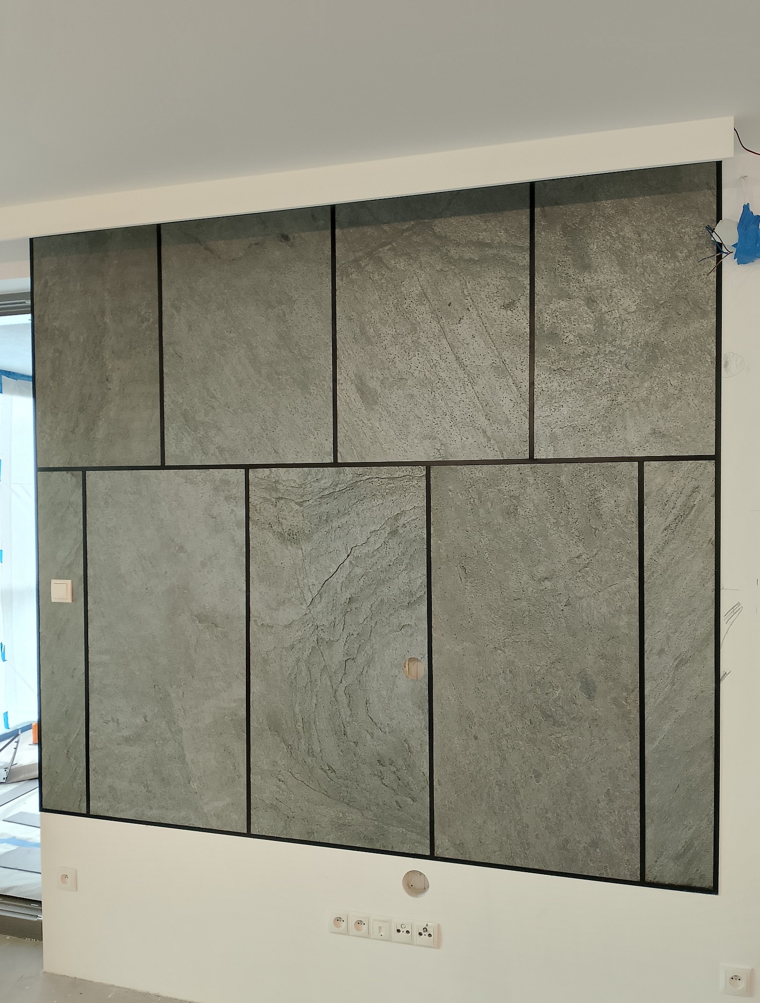 Szara, kamienna okładzina ściany z czarnymi fugami, tworząca geometryczny wzór. Widoczne gniazdka elektryczne i otwór na kabel. Nowoczesne wykończenie wnętrza.