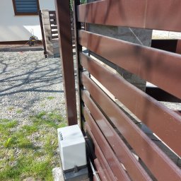 ŁUKASZ STELMACH - Brązowa brama posesji z zamontowanym napędem automatycznym, widoczny z bliska, z domem w tle i fragmentem zieleni.