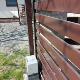 Brązowa brama posesji z zamontowanym napędem automatycznym, widoczny z bliska, z domem w tle i fragmentem zieleni.