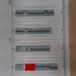 ŁUKASZ STELMACH - Rozdzielnica elektryczna z bezpiecznikami i zabezpieczeniami przeciwprzepięciowymi, widok z bliska na białą obudowę i zainstalowane komponenty.