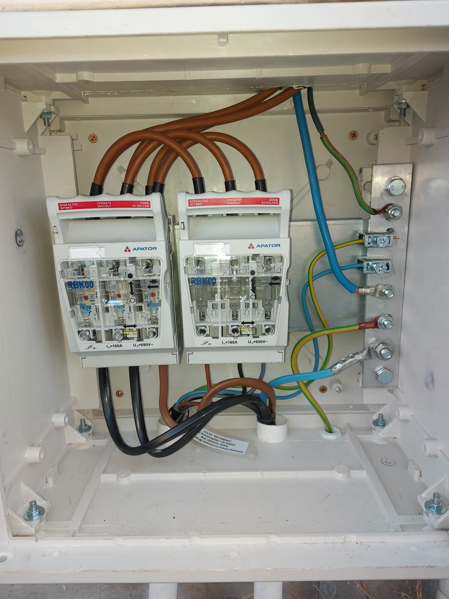 Rozdzielnica elektryczna APATOR RBK00 z bezpiecznikami, widoczne okablowanie w kolorach brązowym, niebieskim i żółto-zielonym. Instalacja w białej skrzynce.
