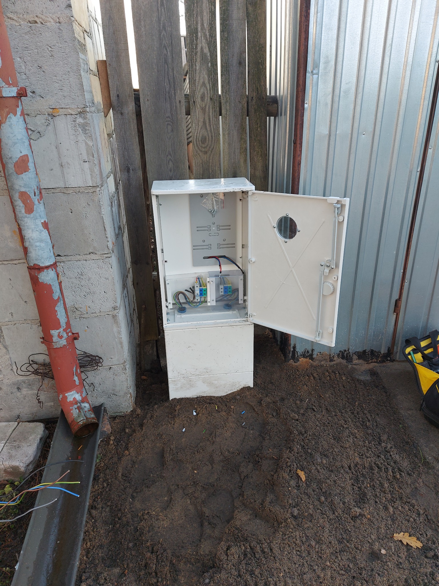 Otwarta, biała skrzynka elektryczna z okrągłym okienkiem, zamontowana na zewnątrz, obok ogrodzenia z blachy falistej i drewnianych desek.