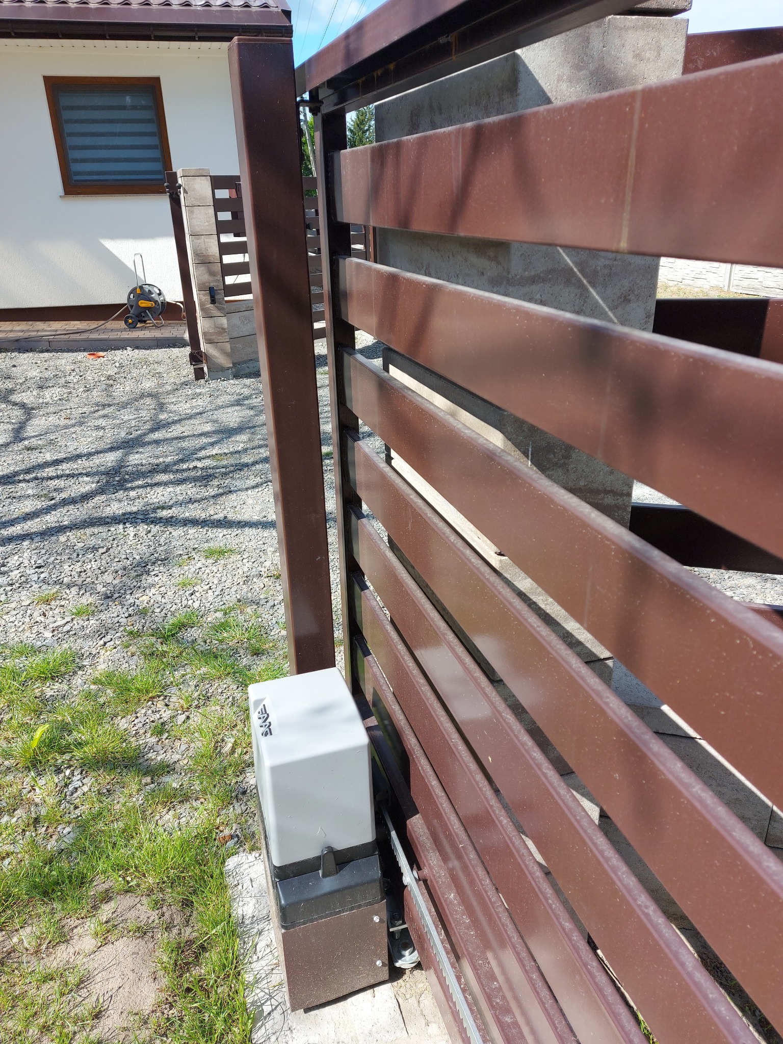 Brązowa brama posesji z zamontowanym napędem automatycznym, widoczny z bliska, z domem w tle i fragmentem zieleni.