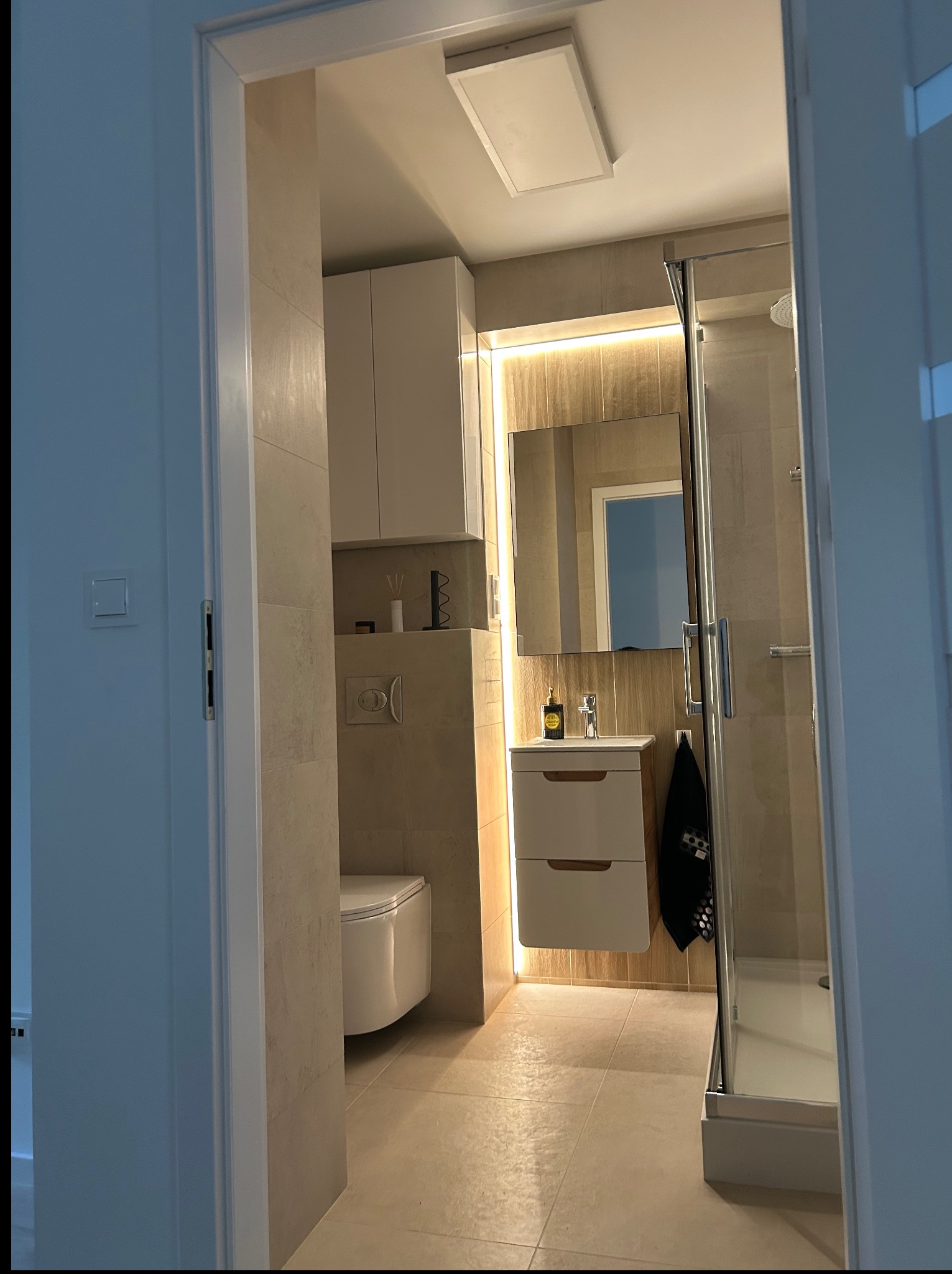 Elegancka łazienka z kabiną prysznicową, umywalką z szafką i toaletą w beżowej tonacji. Oświetlenie LED za lustrem dodaje ciepła. Widok zza uchylonych drzwi.