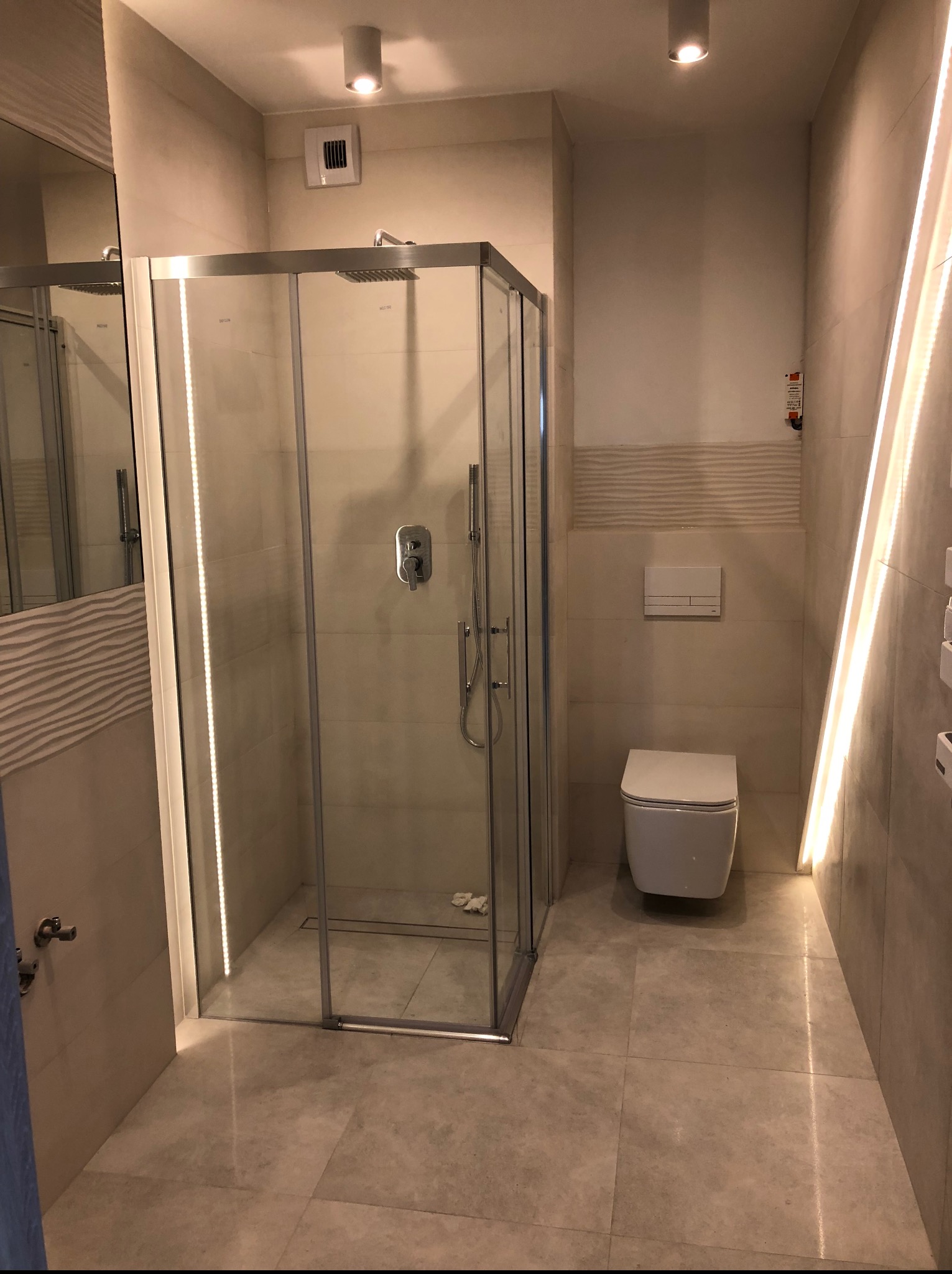 Jasna łazienka z kabiną prysznicową z podświetleniem LED, toaletą i płytkami imitującymi kamień. Widoczny fragment lustra i drzwi.