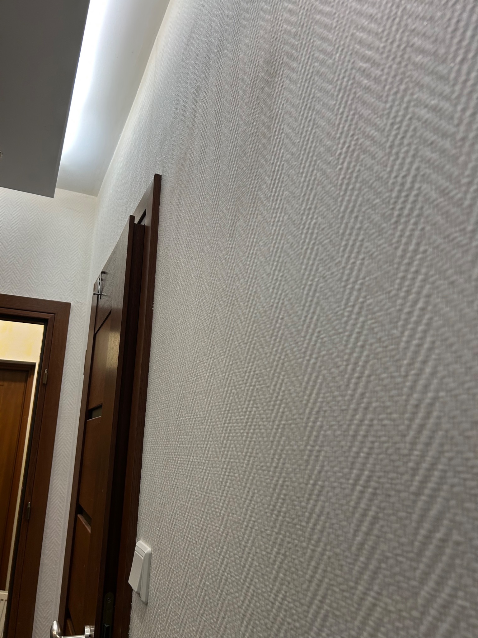 Ściana z tapetą w jodełkę, oświetlenie LED przy suficie, fragment brązowych drzwi. Wykończenie wnętrza mieszkania.