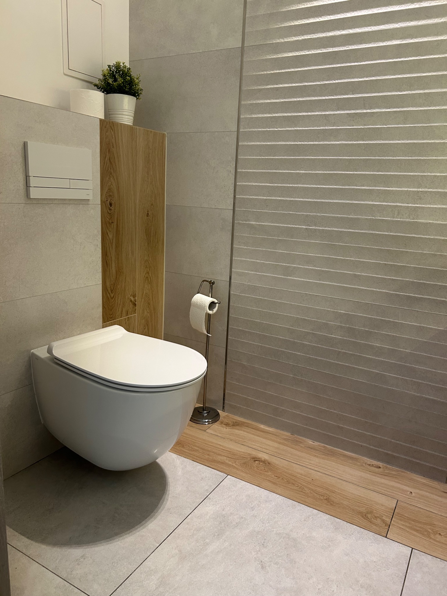 Nowoczesna łazienka z podwieszaną toaletą, szarą płytką i akcentami drewna. Na ścianie dekoracyjna, pozioma faktura. Minimalistyczny styl z rośliną doniczkową.