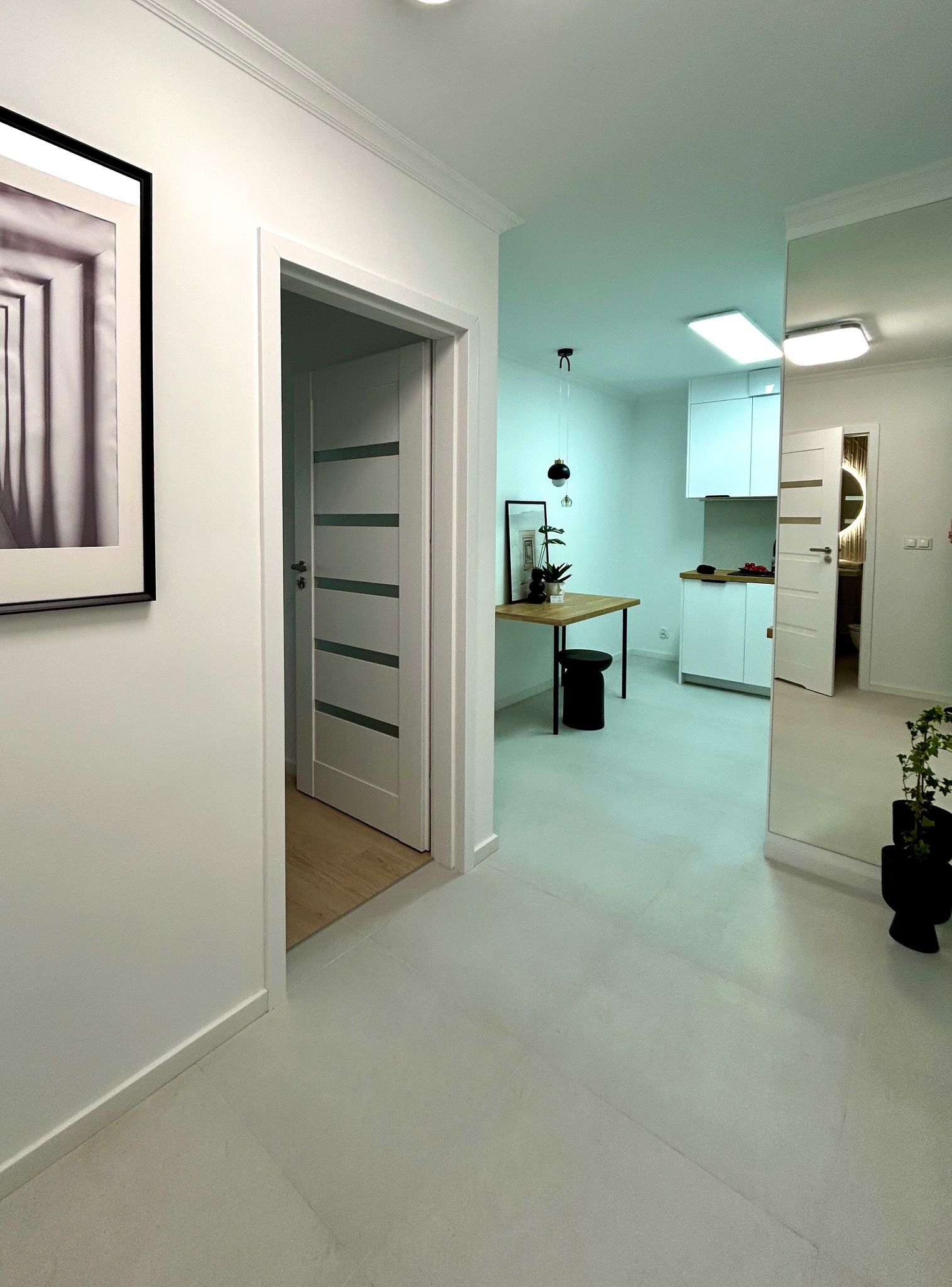 Jasne, minimalistyczne wnętrze mieszkania z otwartą przestrzenią, białymi ścianami, szarą podłogą i drewnianymi akcentami. Widoczne drzwi z przeszkleniami i nowoczesne oświetlenie.