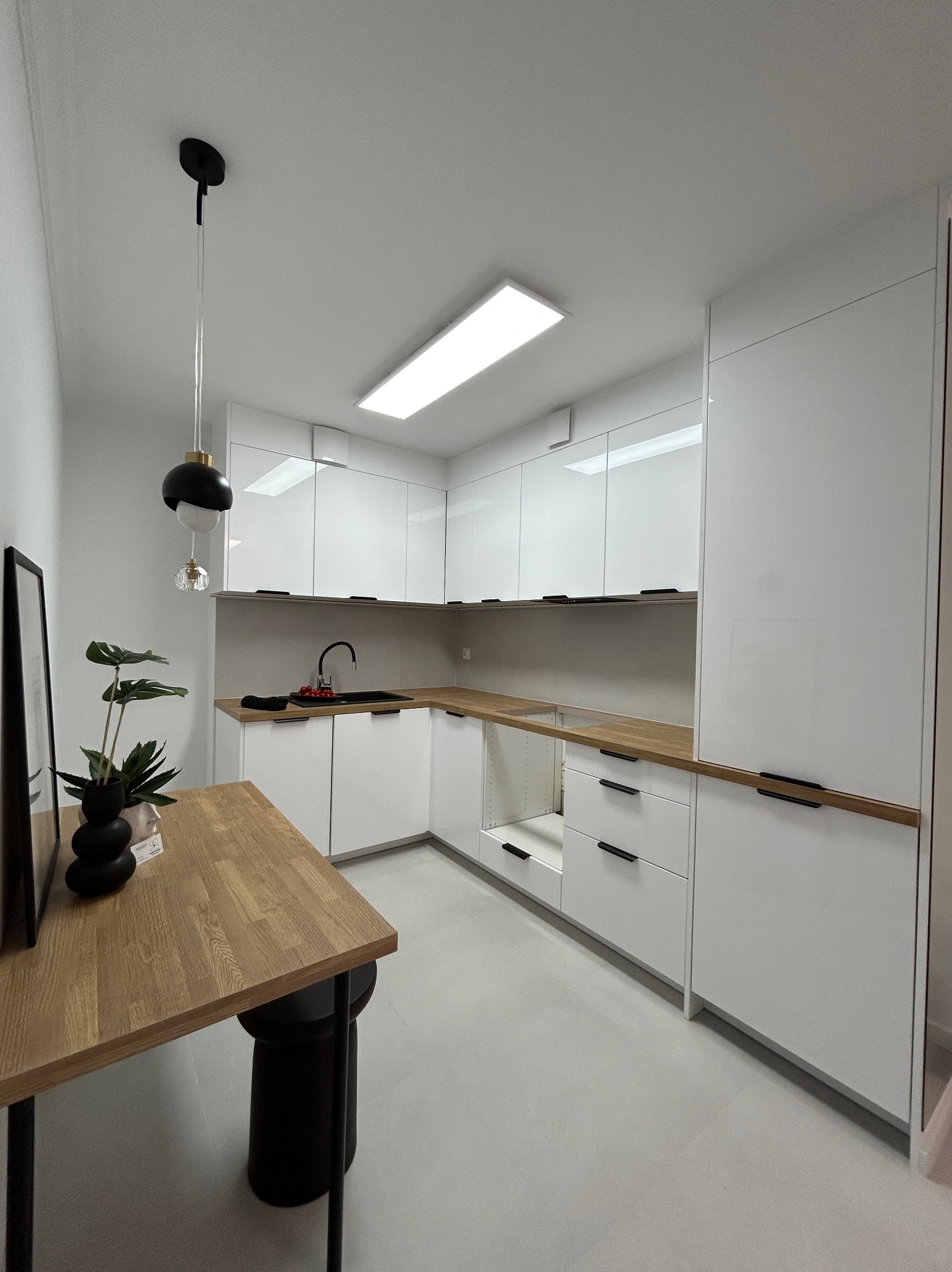 Nowoczesna, minimalistyczna kuchnia z białymi, błyszczącymi frontami szafek i drewnianym blatem. Czarny zlew i bateria. Na pierwszym planie drewniany stół i czarny stołek.