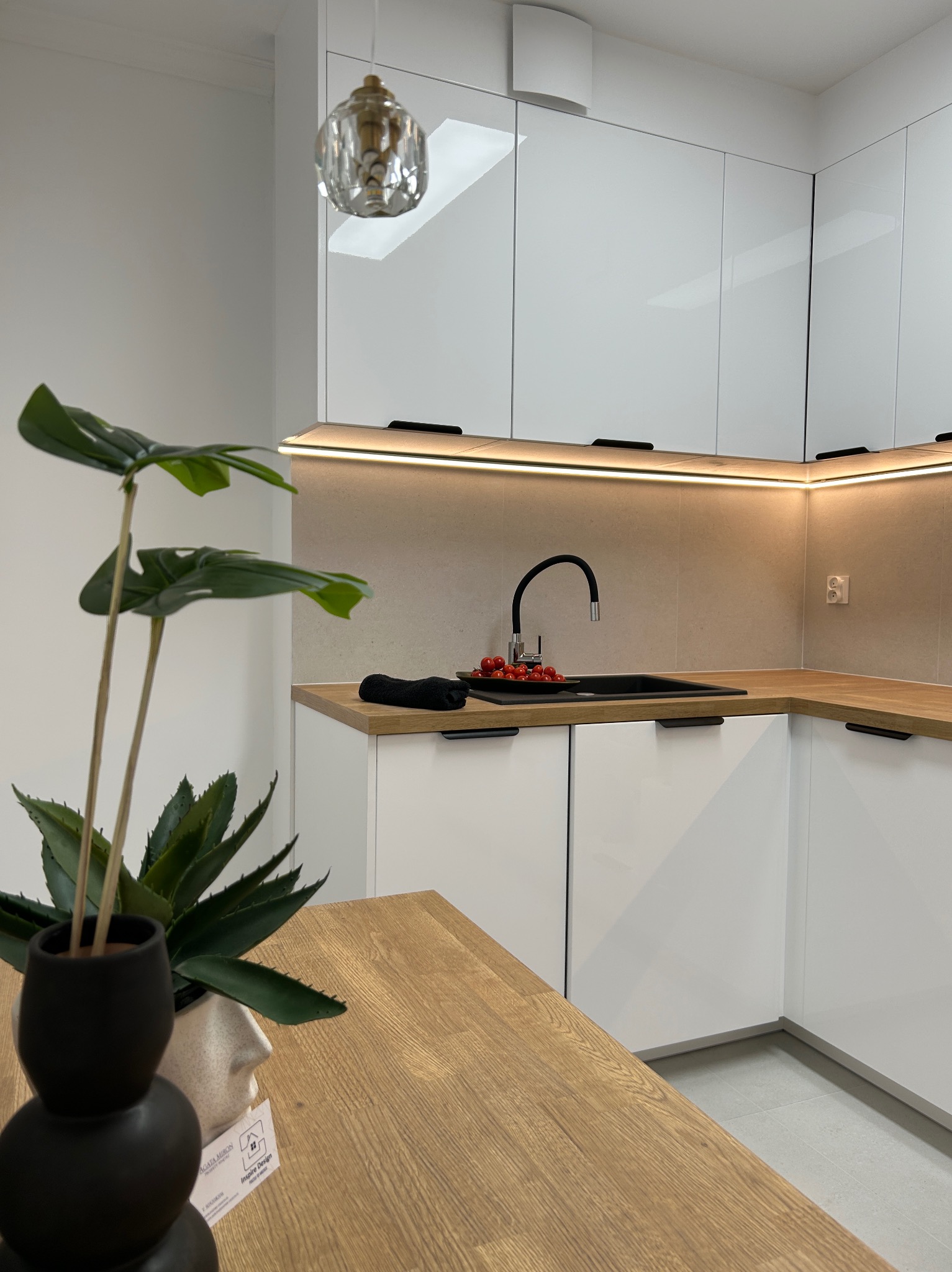 Biała, minimalistyczna kuchnia z drewnianym blatem, czarną armaturą i dekoracyjną rośliną w doniczce. Oświetlenie LED pod szafkami górnymi. Nowoczesny design.