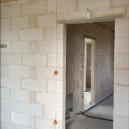 Wnętrze w trakcie budowy, widoczne przewody elektryczne w peszlach i puszki instalacyjne na ścianach z białego bloczka. Surowy betonowy strop, widok na kolejne pomieszczenia.