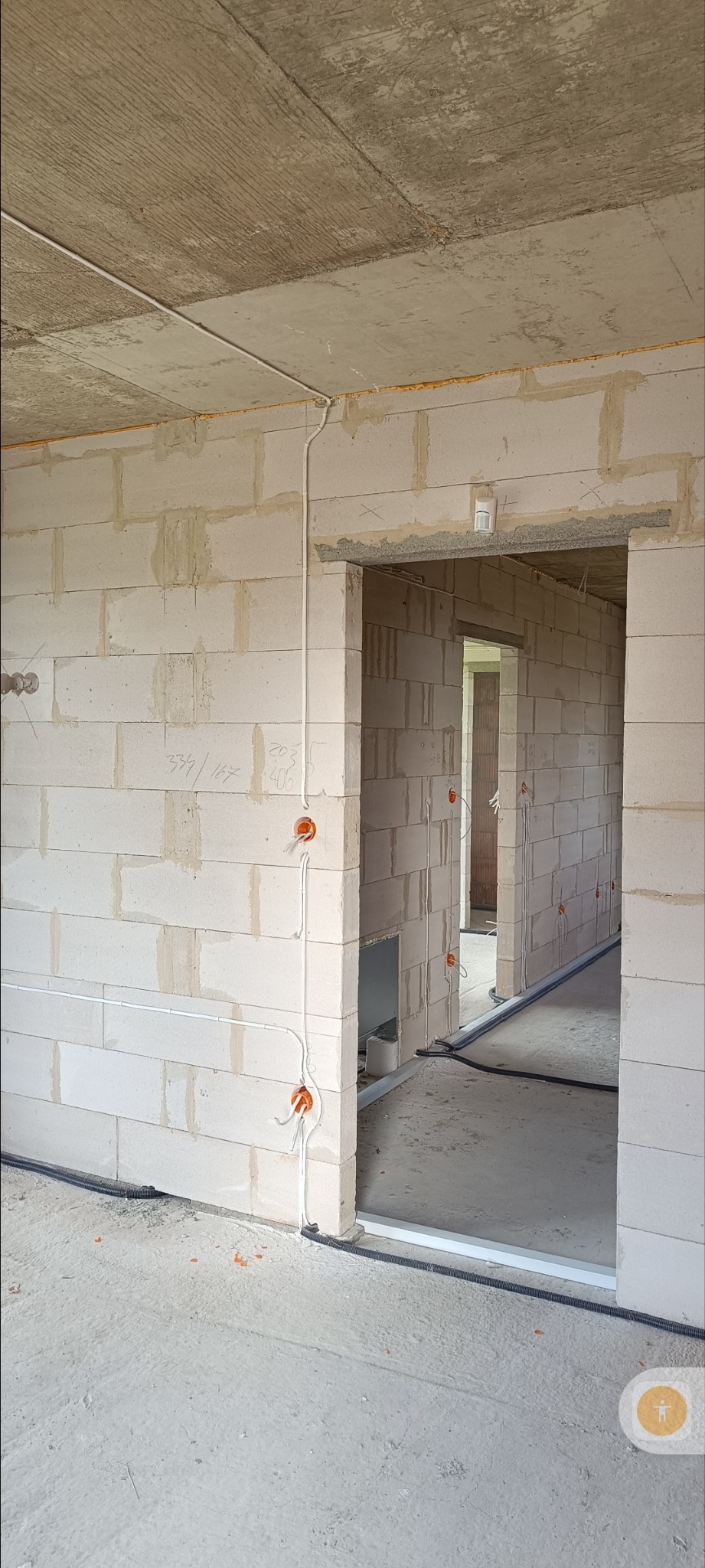 Wnętrze w trakcie budowy, widoczne przewody elektryczne w peszlach i puszki instalacyjne na ścianach z białego bloczka. Surowy betonowy strop, widok na kolejne pomieszczenia.