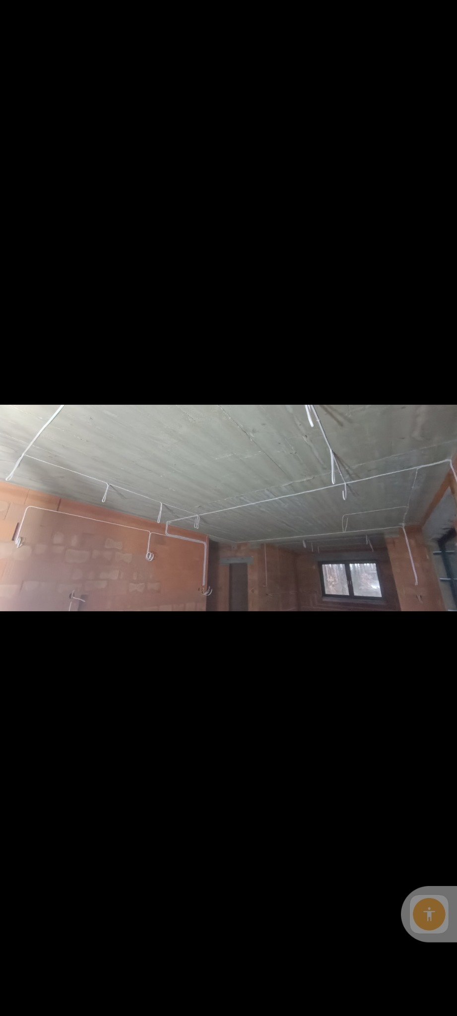 Wnętrze w stanie surowym z rozprowadzonymi kablami elektrycznymi pod sufitem i na ścianach z cegły. Widoczne okno i otwór drzwiowy.