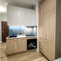 Tobimeble- Meble na wymiar - Kompaktowa, jasna kuchnia na wymiar w mieszkaniu: białe szafki wiszące, drewnopodobne dolne szafki z czarnym zlewem i piekarnikiem, wysoka zabudowa.