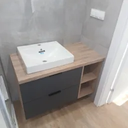 Szara szafka łazienkowa z drewnianym blatem i białą umywalką Roca. Dwie szuflady i półki po prawej. Całość na tle szarych płytek i jasnej podłogi.