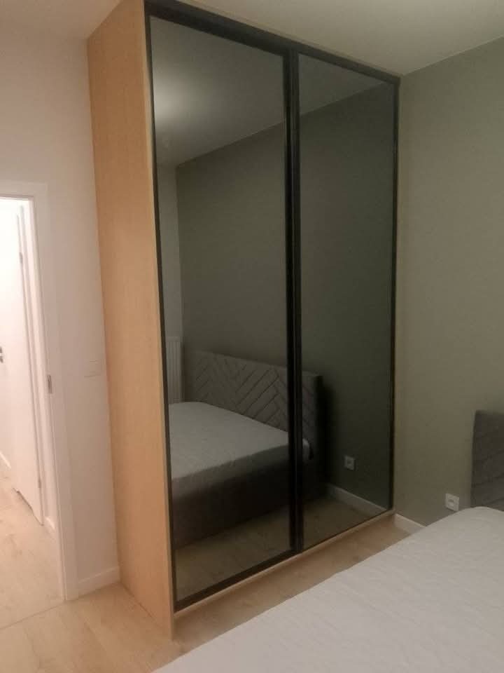 Szafa wnękowa z przesuwnymi drzwiami lustrzanymi i czarnymi ramami, widoczna w sypialni z elementami odbicia wnętrza pokoju. Jasna okleina po bokach.