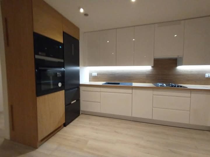 Nowoczesna kuchnia: białe szafki z połyskiem, drewniany akcent w zabudowie piekarnika i lodówki, podświetlany blat roboczy, jasna podłoga.