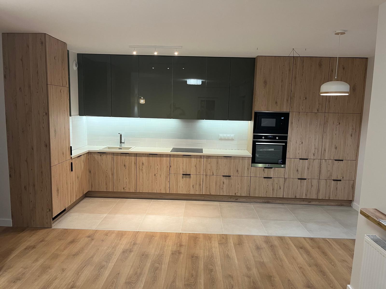 Nowoczesna kuchnia z drewnianymi frontami i czarnymi szafkami wiszącymi, oświetlona lampami punktowymi i wiszącą lampą. Widok na całe pomieszczenie, parkiet i płytki.