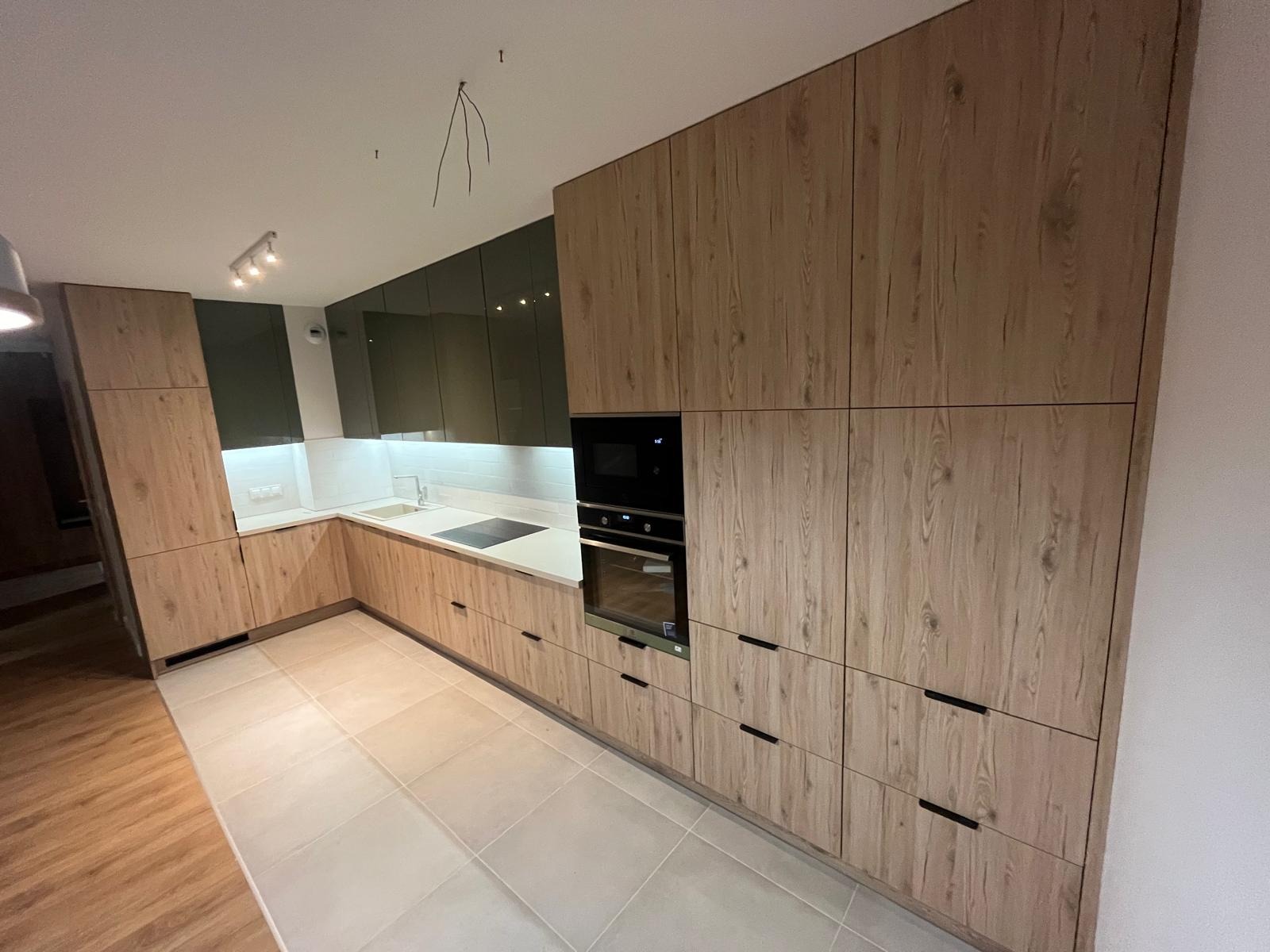 Nowoczesna kuchnia z drewnianymi frontami i zielonymi szafkami wiszącymi. Minimalistyczne uchwyty, jasny blat i płytki podłogowe. Widoczne przewody elektryczne.