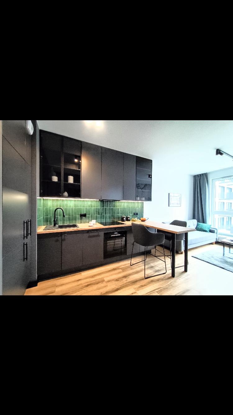 Minimalistyczna kuchnia w odcieniach szarości z drewnianym blatem i zieloną mozaiką na ścianie. Uwagę przykuwa nowoczesne oświetlenie LED pod szafkami.