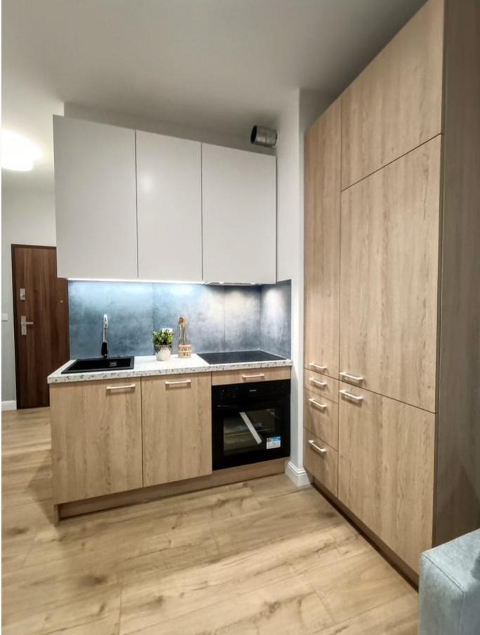 Kompaktowa, jasna kuchnia na wymiar w mieszkaniu: białe szafki wiszące, drewnopodobne dolne szafki z czarnym zlewem i piekarnikiem, wysoka zabudowa.