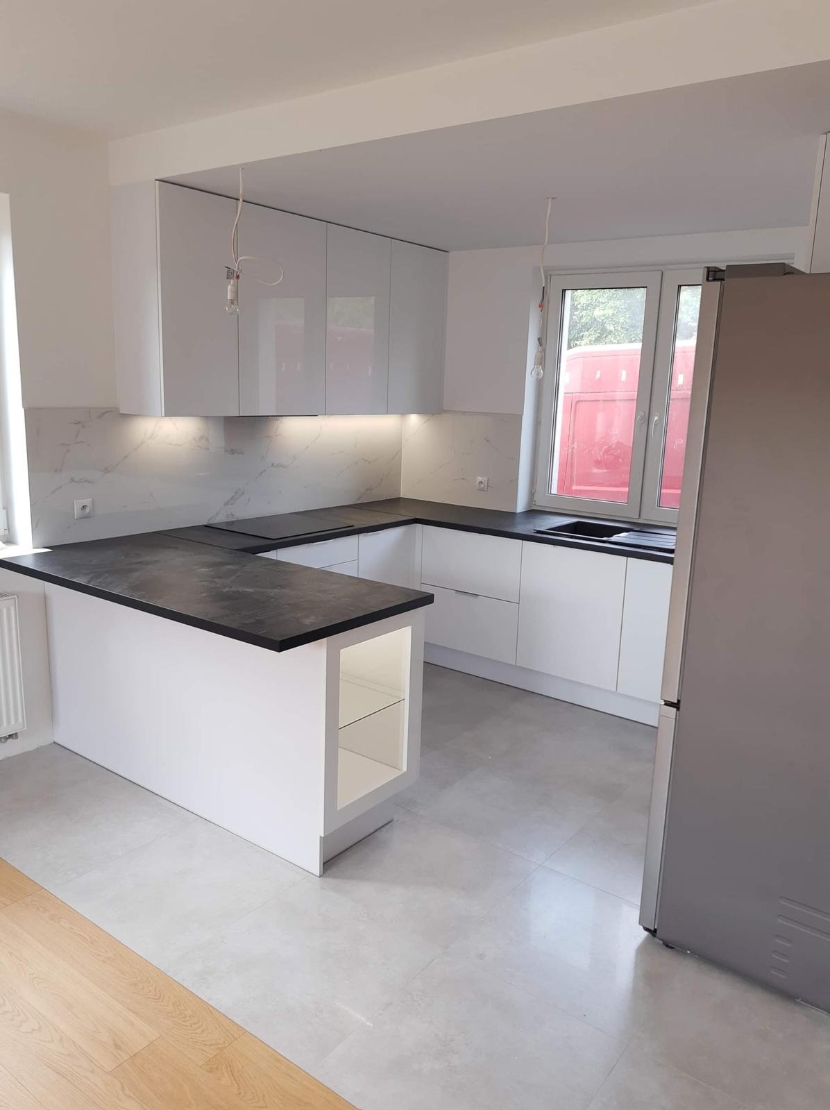 Nowoczesna, biała kuchnia z czarnym blatem i wyspą. Błyszczące fronty szafek, marmurowy backsplash, minimalistyczne oświetlenie. Widok na okno i fragment lodówki.