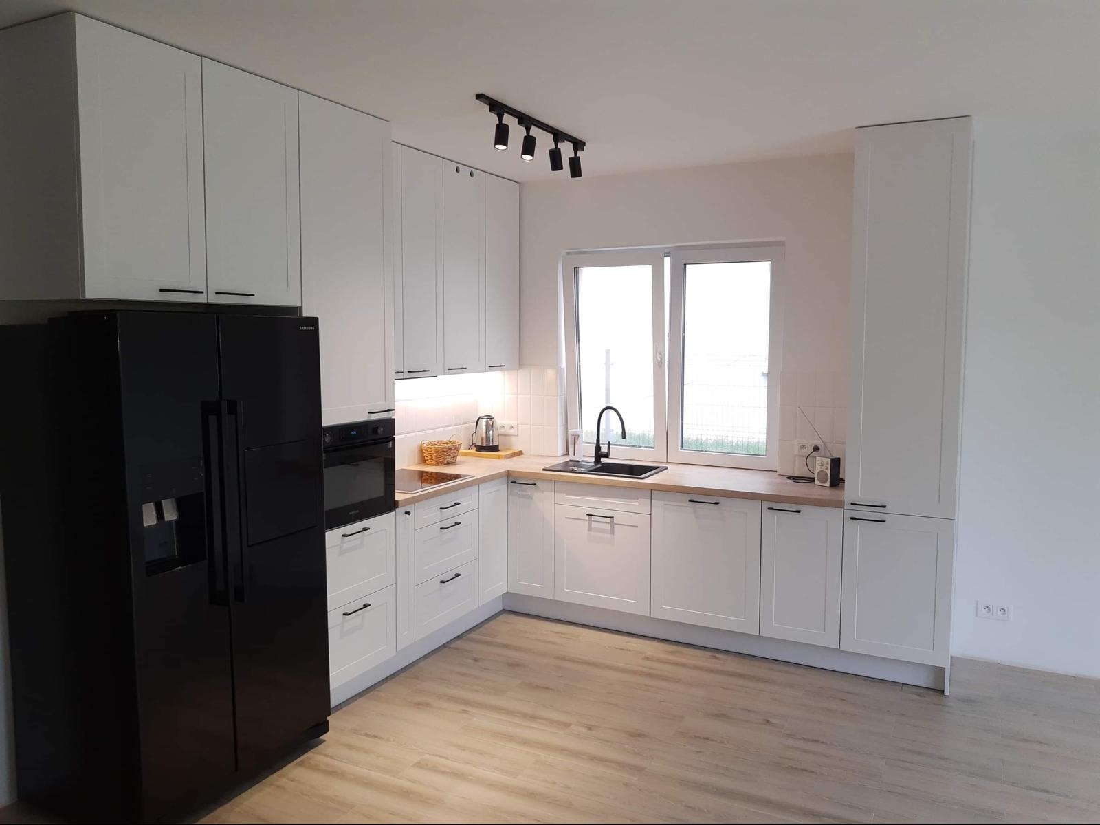 Biała kuchnia z czarnymi akcentami: nowoczesne szafki z uchwytami, czarny zlew i lodówka, drewniany blat, oświetlenie punktowe. Minimalistyczny design.