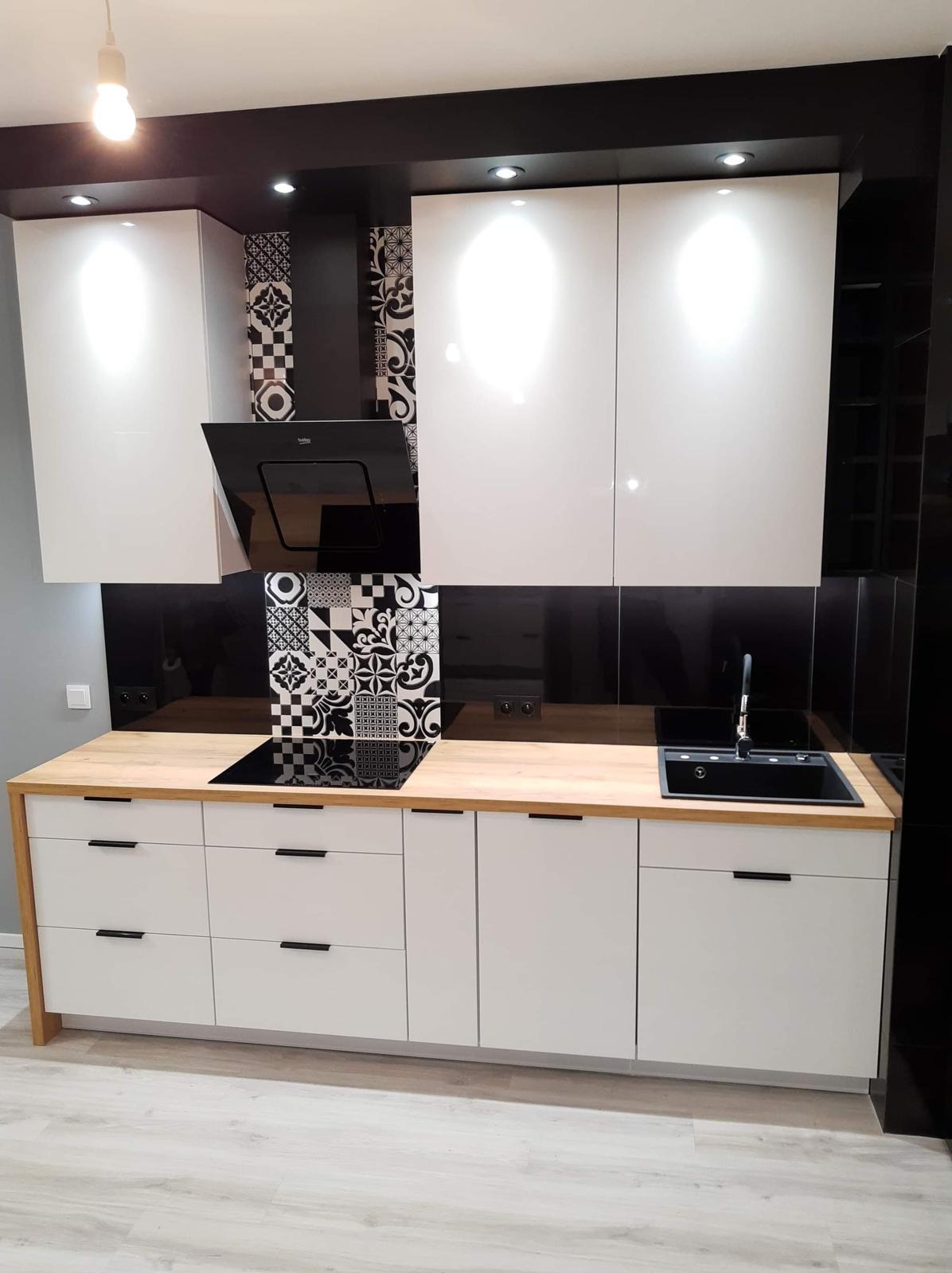 Nowoczesna kuchnia: białe szafki z czarnymi uchwytami, drewniany blat i dekoracyjne płytki nad indukcją. Czarny okap i szklany panel dodają elegancji.