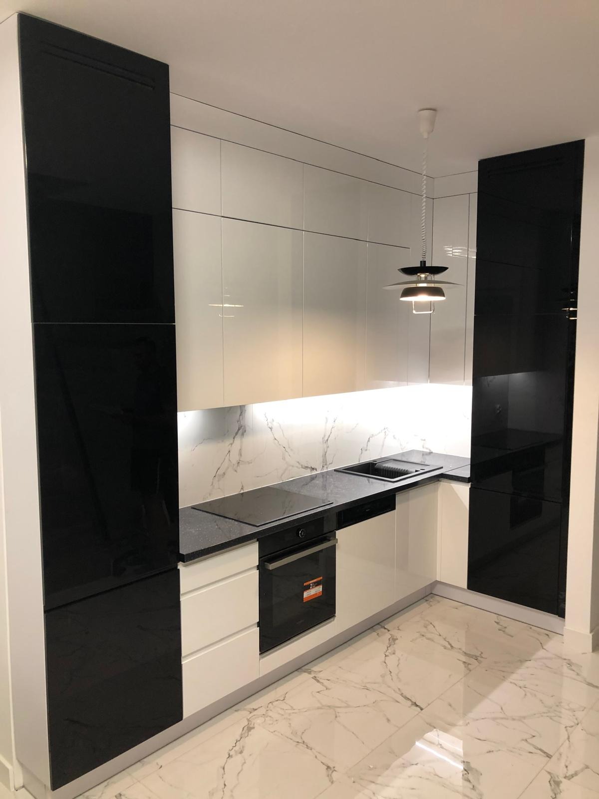 Nowoczesna, narożna kuchnia z białymi i czarnymi, lakierowanymi frontami. Blat z ciemnego kamienia, marmurowa ściana, designerska lampa. Minimalistyczny styl.