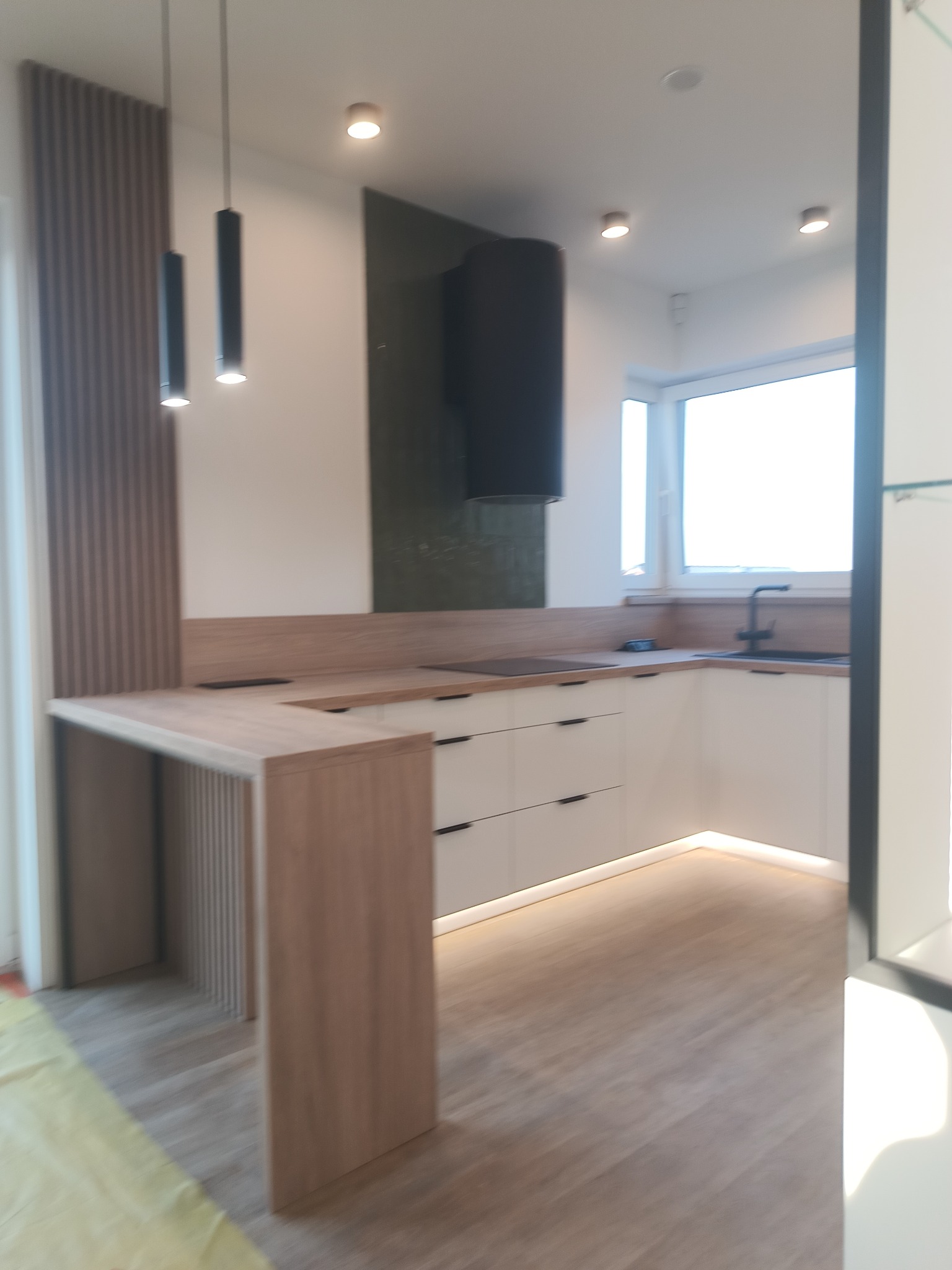 Nowoczesna, jasna kuchnia z wyspą i oświetleniem LED pod szafkami. Blat w kolorze drewna, czarny okap i minimalistyczne lampy wiszące.