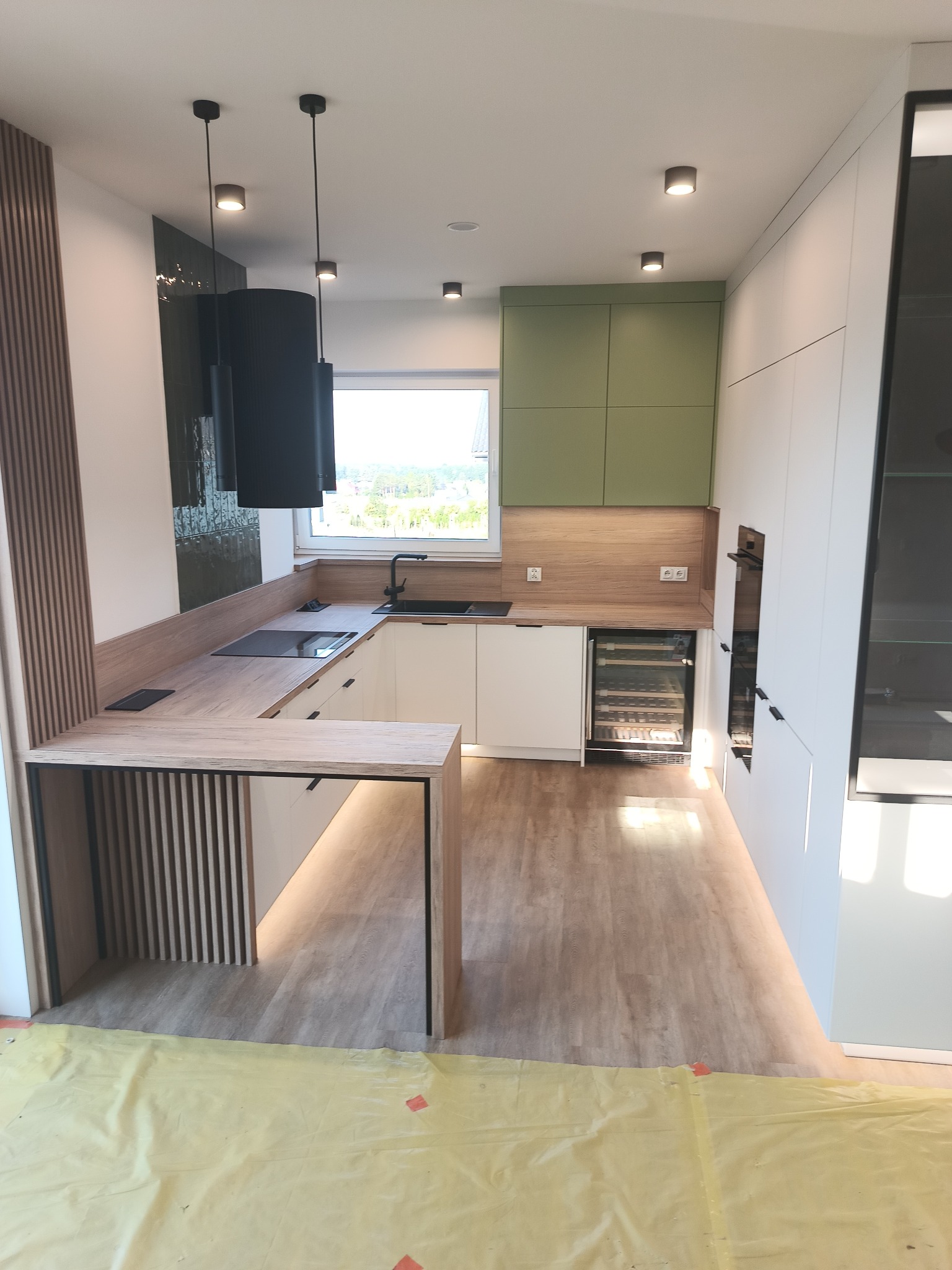 Nowoczesna kuchnia z wyspą i barkiem, białe fronty, drewniany blat, zielone szafki wiszące, czarne lampy wiszące, podłoga imitująca drewno.