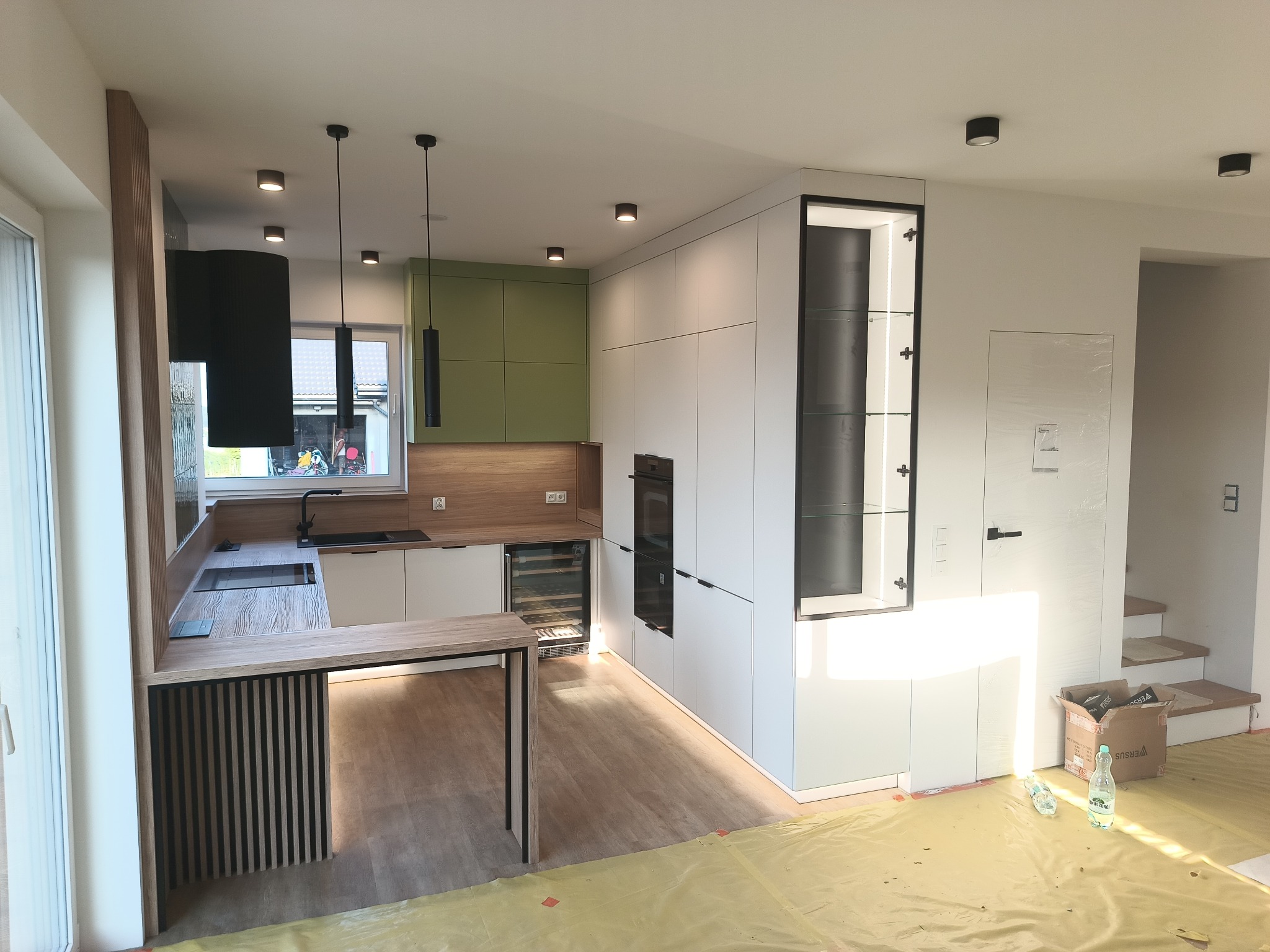 Nowoczesna kuchnia z wyspą i zabudową w bieli oraz akcentami drewna i zieleni, Radomsko. Widoczne oświetlenie podszafkowe i sprzęt AGD w zabudowie.