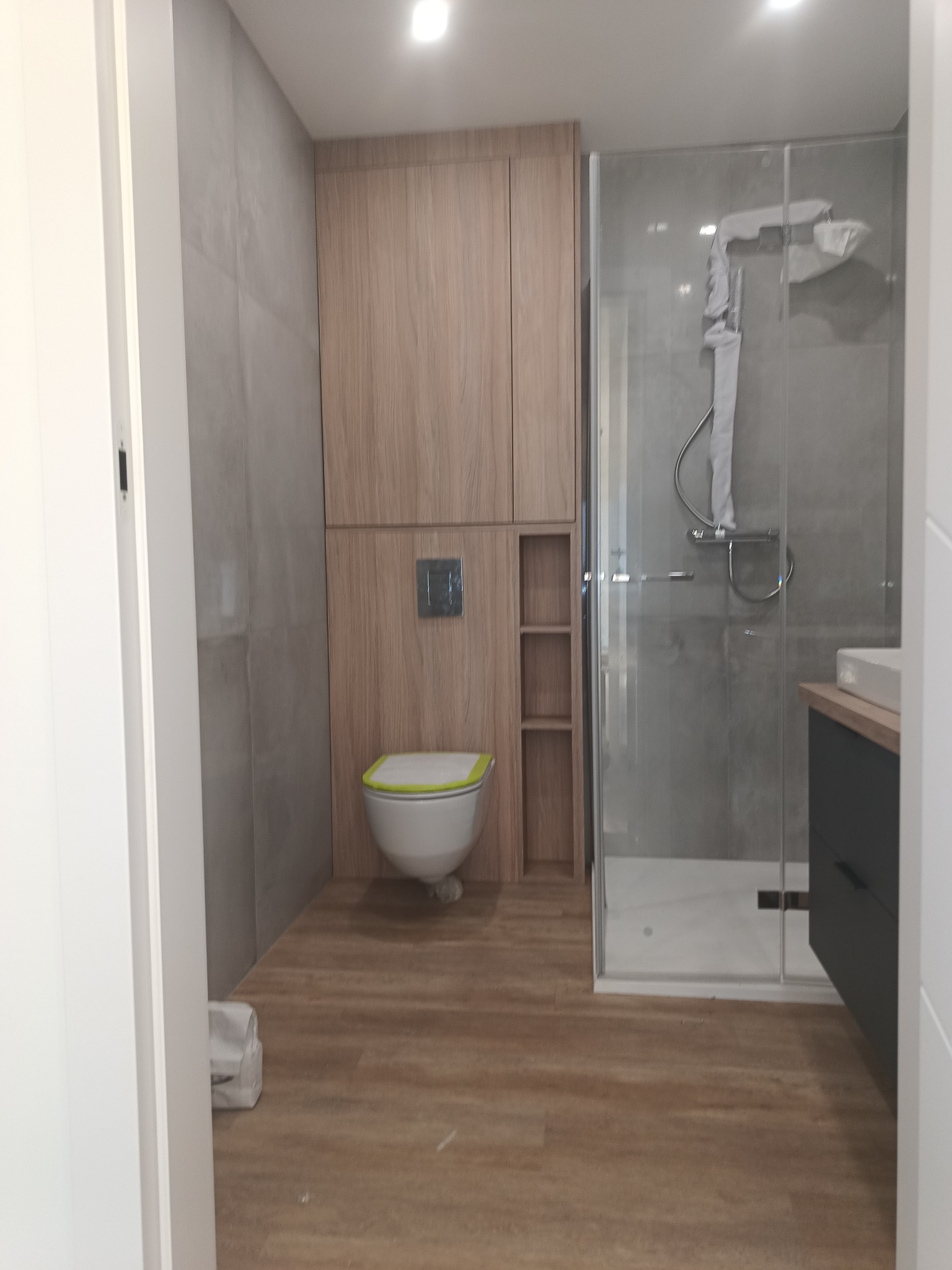 Nowoczesna łazienka z drewnianą zabudową nad toaletą i kabiną prysznicową. Umywalka z szafką w ciemnym kolorze. Podłoga imitująca drewno. Minimalistyczny design.