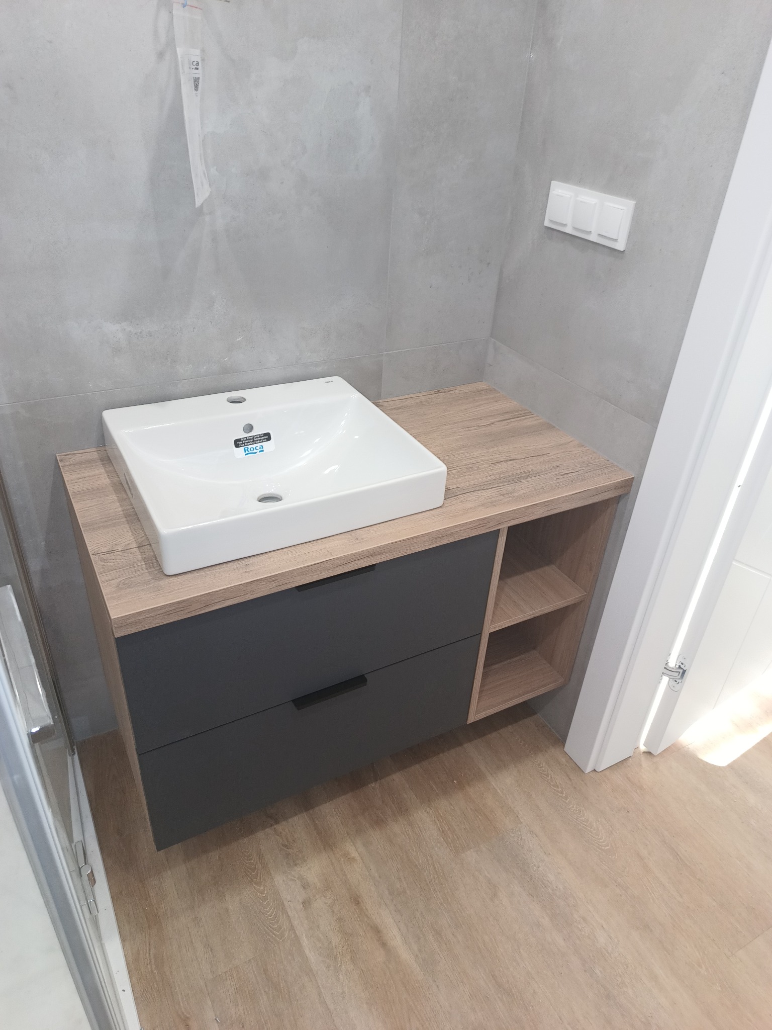 Szara szafka łazienkowa z drewnianym blatem i białą umywalką Roca. Dwie szuflady i półki po prawej. Całość na tle szarych płytek i jasnej podłogi.