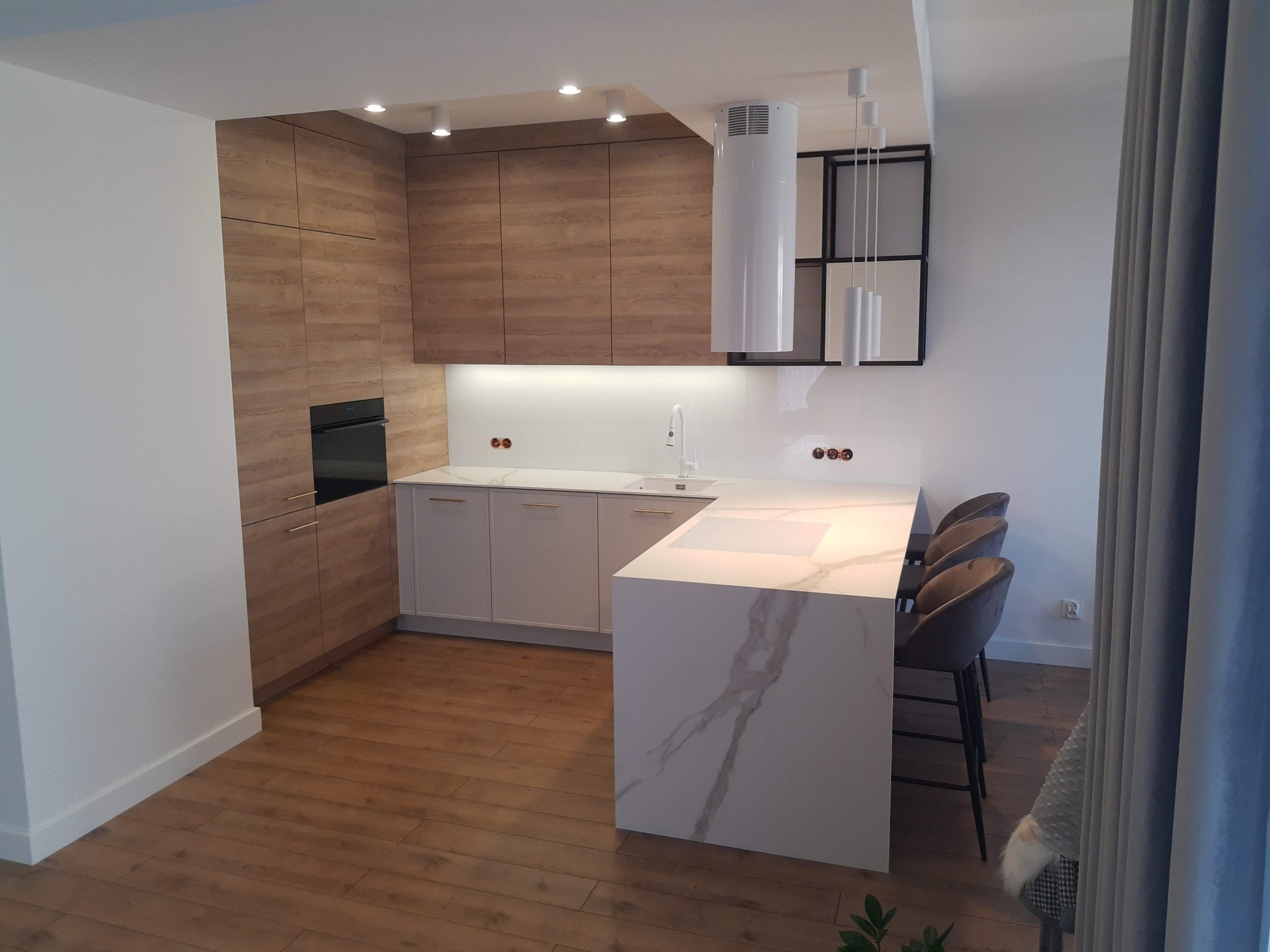 Nowoczesna kuchnia z drewnianymi szafkami górnymi i wyspą z białego marmuru. Uwagę przykuwa minimalistyczny okap i designerskie lampy wiszące.