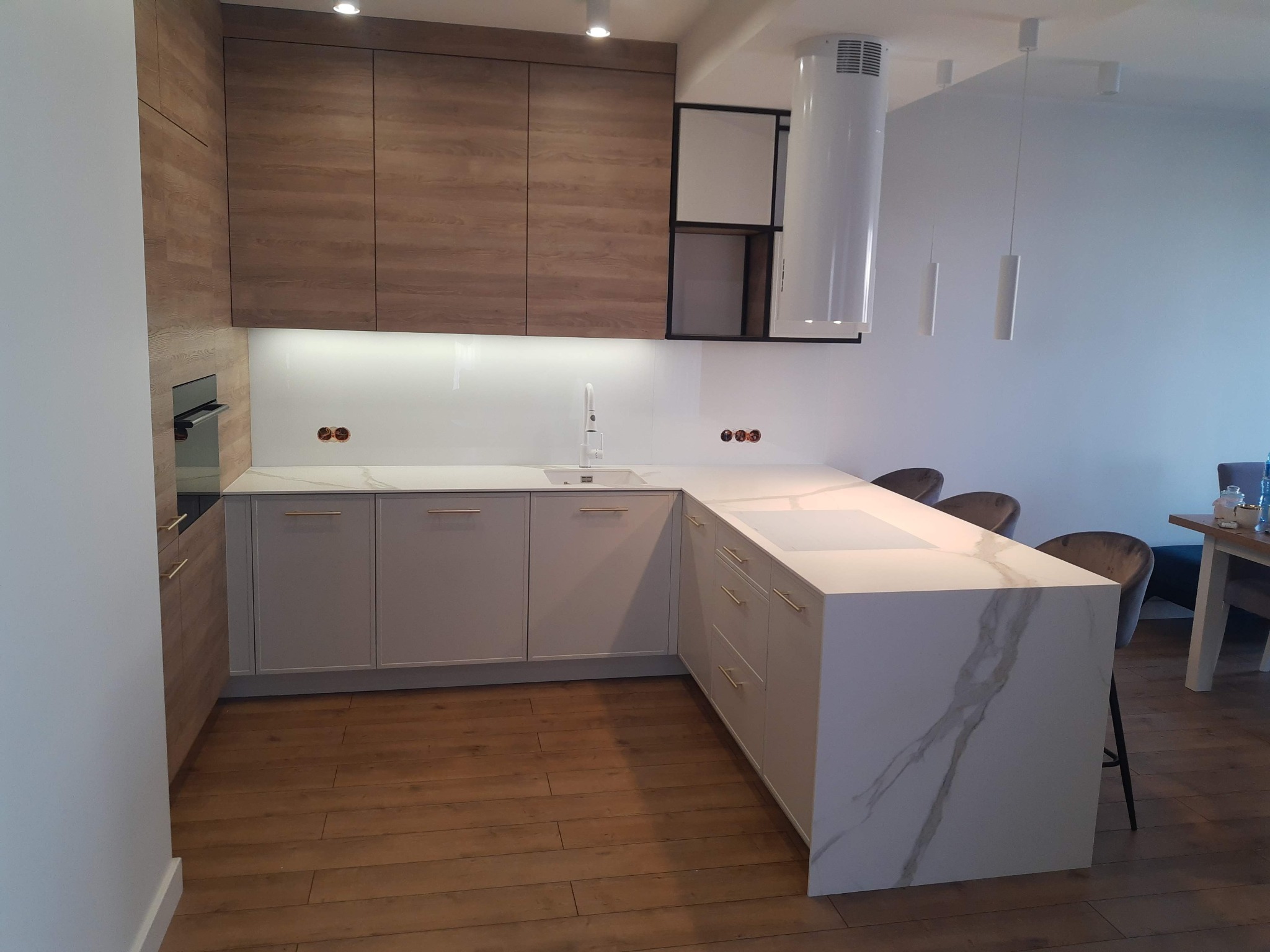 Nowoczesna kuchnia z drewnianymi szafkami górnymi i białymi dolnymi, wyspa z blatem imitującym marmur, oświetlenie punktowe i drewniana podłoga.